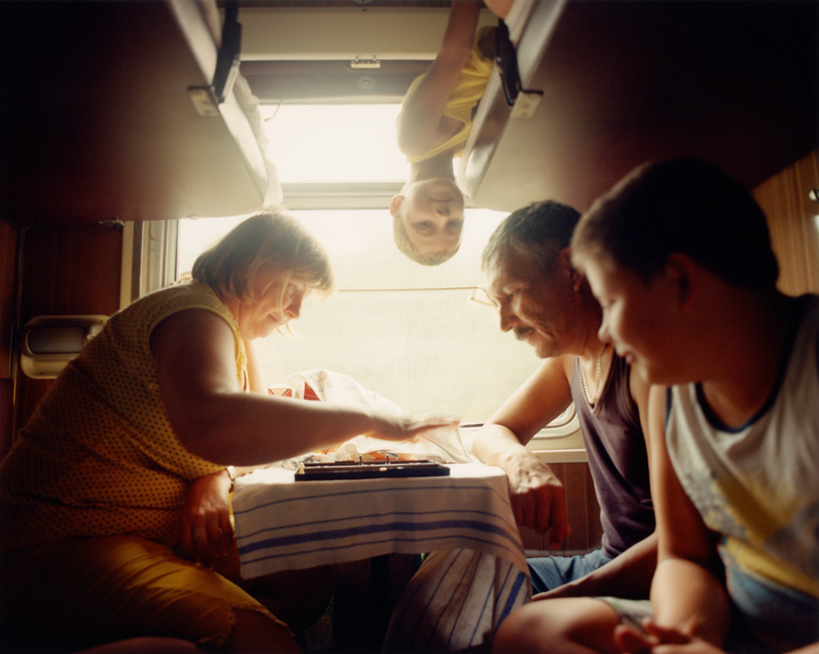 Inside a sunlit camper van or train, an older woman and man gather at a small table while a child hangs upside down from the ceiling, peering towards the camera.